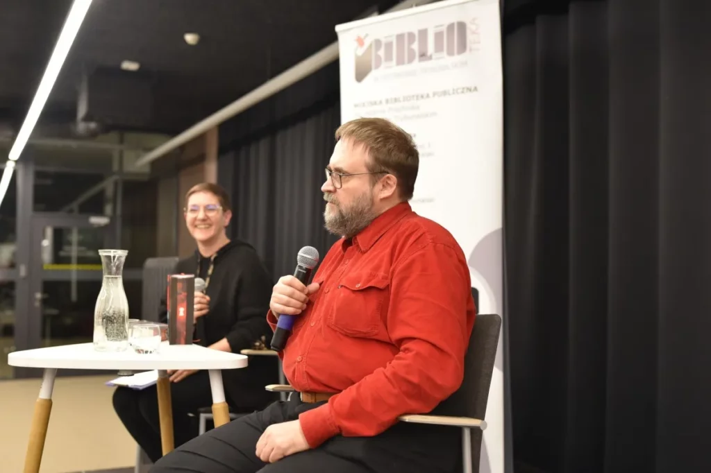 Andrzej Pilipiuk siedzi przy stoliku i trzyma w dłoni mikrofon, odpowiadając na pytania podczas spotkania autorskiego. Obok siedzi prowadząca wydarzenie, która uśmiecha się i również trzyma mikrofon. Na stoliku stoją książki autora, karafka z wodą oraz szklanka. W tle widoczny jest roll-up Miejskiej Biblioteki Publicznej w Piotrkowie Trybunalskim.