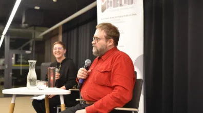 Andrzej Pilipiuk siedzi przy stoliku i trzyma w dłoni mikrofon, odpowiadając na pytania podczas spotkania autorskiego. Obok siedzi prowadząca wydarzenie, która uśmiecha się i również trzyma mikrofon. Na stoliku stoją książki autora, karafka z wodą oraz szklanka. W tle widoczny jest roll-up Miejskiej Biblioteki Publicznej w Piotrkowie Trybunalskim.