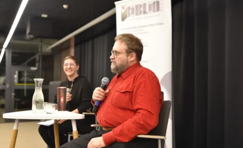 Andrzej Pilipiuk siedzi przy stoliku i trzyma w dłoni mikrofon, odpowiadając na pytania podczas spotkania autorskiego. Obok siedzi prowadząca wydarzenie, która uśmiecha się i również trzyma mikrofon. Na stoliku stoją książki autora, karafka z wodą oraz szklanka. W tle widoczny jest roll-up Miejskiej Biblioteki Publicznej w Piotrkowie Trybunalskim.