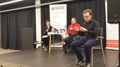 Prowadząca spotkanie siedzi na scenie i trzyma mikrofon, zadając pytanie gościowi. Obok niej siedzi Andrzej Pilipiuk, który odpowiada na pytania podczas spotkania autorskiego. Z prawej strony siedzi tłumacz języka migowego, który przekłada wypowiedzi na Polski Język Migowy. Na stoliku znajdują się książki autora, karafka z wodą oraz szklanka.