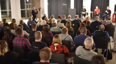 Publiczność siedzi w rzędach krzeseł i słucha spotkania autorskiego. Na scenie Andrzej Pilipiuk rozmawia z prowadzącą spotkanie. Obok obecny jest tłumacz języka migowego przekładający wypowiedzi dla osób niesłyszących. Z boku sali operator kamery nagrywa przebieg wydarzenia.