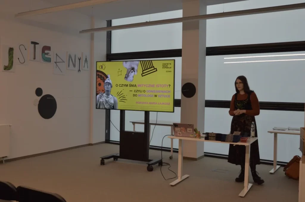 Zdjęcie przedstawia prelekcję w Strefie SOWA, podczas której prowadząca stoi obok ekranu z prezentacją zatytułowaną „O czym śnią mityczne istoty?”, a przed nią znajduje się stolik z laptopem i materiałami, w jasnej i nowoczesnej przestrzeni edukacyjnej.