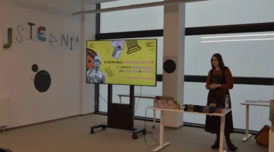 Zdjęcie przedstawia prelekcję w Strefie SOWA, podczas której prowadząca stoi obok ekranu z prezentacją zatytułowaną „O czym śnią mityczne istoty?”, a przed nią znajduje się stolik z laptopem i materiałami, w jasnej i nowoczesnej przestrzeni edukacyjnej.