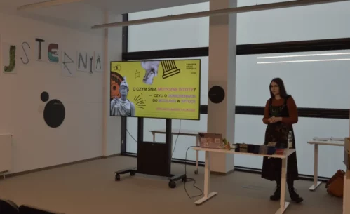 Zdjęcie przedstawia prelekcję w Strefie SOWA, podczas której prowadząca stoi obok ekranu z prezentacją zatytułowaną „O czym śnią mityczne istoty?”, a przed nią znajduje się stolik z laptopem i materiałami, w jasnej i nowoczesnej przestrzeni edukacyjnej.