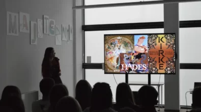 Zdjęcie przedstawia prelekcję w Strefie SOWA, podczas której prowadząca stoi obok ekranu z prezentacją dotyczącą mitologii w kulturze współczesnej, a na slajdzie widoczne są odniesienia do postaci Hadesa oraz książki „Kirke” Madeline Miller, podczas gdy publiczność siedzi i uważnie słucha wystąpienia.