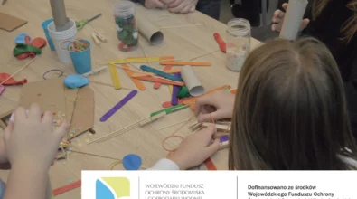 Na zdjęciu widoczny jest stół warsztatowy, przy którym dzieci wspólnie tworzą instrumenty muzyczne z materiałów z recyklingu, takich jak patyczki, rolki po papierze, gumki recepturki, balony i plastikowe zakrętki. Fotografia dokumentuje kreatywne zajęcia edukacyjne realizowane w ramach zadania EkoTEKA, dofinansowanego ze środków Wojewódzkiego Funduszu Ochrony Środowiska i Gospodarki Wodnej w Łodzi.