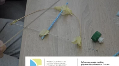 Na zdjęciu widoczny jest fragment stołu warsztatowego, na którym leżą elementy tworzonego instrumentu z materiałów recyklingowych: słomka, sznurek, patyczki oraz zakrętka od butelki połączone taśmą. Fotografia dokumentuje zajęcia edukacyjne realizowane w ramach projektu EkoTEKA, dofinansowanego ze środków Wojewódzkiego Funduszu Ochrony Środowiska i Gospodarki Wodnej w Łodzi.