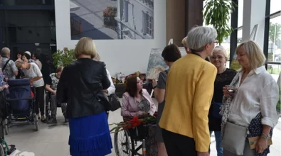 Zakończenie wernisażu w Mediatece 800-lecia. W przestronnym, jasnym holu goście rozmawiają i gratulują artystce, która siedzi na wózku inwalidzkim, trzymając bukiet czerwonych kwiatów. Kobieta w żółtej marynarce i srebrnowłosa dama w białej koszuli prowadzą ożywioną rozmowę. W tle inni uczestnicy wydarzenia, osoby na wózkach, wystawione na sztalugach obrazy oraz duża fotografia architektoniczna na ścianie. Przez przeszklone ściany wpada naturalne światło, tworząc przyjazną atmosferę. W lewym dolnym rogu widoczne logo Mediateki.