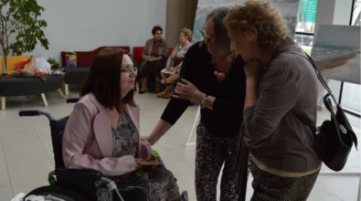 Artystka na wózku rozmawia z dwiema kobietami podczas wernisażu w Mediatece 800-lecia. Atmosfera pełna serdeczności i wzruszenia, w tle obrazy oraz inni goście wydarzenia. Na ławkach widoczne są bukiety kwiatów, symboliczne podziękowania dla twórczyni za jej dzieła.