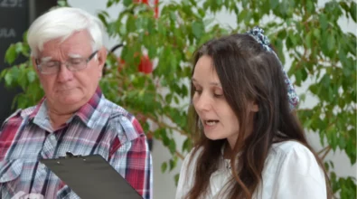 Zbliżenie na moment przemówienia podczas wernisażu w Mediatece 800-lecia. Kobieta w białej bluzce i ozdobnej opasce na włosach odczytuje treść z czarnej teczki, z zaangażowaniem wypowiadając słowa. Obok niej starszy mężczyzna w koszuli w kratę uważnie słucha, spoglądając w dół. W tle zielona roślina doniczkowa oraz fragment nowoczesnego wnętrza Mediateki. W lewym dolnym rogu widoczne logo Mediateki.