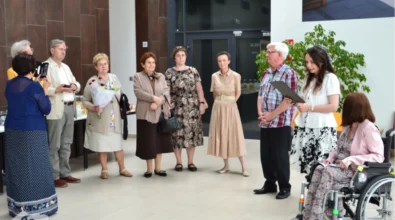 Uroczyste zakończenie wernisażu w Mediatece 800-lecia. Kobieta w białej bluzce i czarno-białej spódnicy odczytuje tekst z teczki, stojąc obok starszego mężczyzny w koszuli w kratę. Przed nimi grupa elegancko ubranych osób, w tym kobieta trzymająca bukiet czerwonych róż. Po prawej stronie artystka, siedząca na wózku inwalidzkim, słucha z uśmiechem. W tle stolik z poczęstunkiem, przeszklone wejście do auli oraz zielone rośliny dekoracyjne. W lewym dolnym rogu widoczne logo Mediateki.