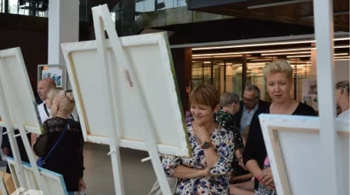 Dwie kobiety stojące przed sztalugami z obrazami na wystawie w Mediatece 800-lecia. Jedna z nich uśmiecha się, spoglądając na dzieło, druga z zainteresowaniem przygląda się szczegółom. W tle pozostali uczestnicy wernisażu, w tym osoby oglądające prace i prowadzące rozmowy. Nowoczesne wnętrze z przeszklonymi ścianami wypełnia ciepłe światło, podkreślając atmosferę kulturalnego wydarzenia.