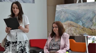 Uroczyste zakończenie wernisażu w Mediatece 800-lecia. Kobieta w białej bluzce i spódnicy w czarno-biały wzór odczytuje tekst z teczki, stojąc obok artystki siedzącej na wózku inwalidzkim. Malarka, ubrana w wzorzystą sukienkę i jasnoróżową marynarkę, z uśmiechem słucha wystąpienia. W tle obraz przedstawiający górski krajobraz oraz nowoczesne wnętrze Mediateki z kolorowymi siedziskami.