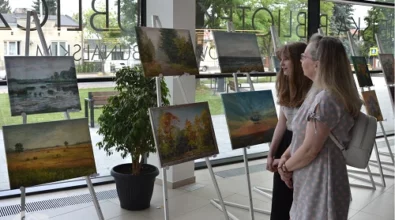 Fotografia przedstawiająca kobietę stojącą obok dziewczynki. Przed nimi obrazy autorstwa Fabiola Mikler. W tle drzewo w doniczce, szklane ściany, główne wejście Mediateki. Zdjęcie własnością MBP Piotrków.