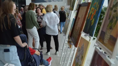 .Wernisaż wystawy malarskiej w Mediatece 800-lecia. Wzdłuż jasnej, przeszklonej przestrzeni ustawione są sztalugi z kolorowymi obrazami, głównie o tematyce kwiatowej i pejzażowej. W tle liczni goście, rozmawiający w grupach i podziwiający prace. Po lewej stronie kobieta w okularach popycha wózek inwalidzki, w którym siedzi dziecko ubrane w czerwony strój i sportowe buty. W lewym dolnym rogu logo Mediateki.