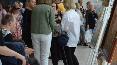 Wernisaż wystawy malarskiej w Mediatece 800-lecia. Wzdłuż jasnej, przeszklonej przestrzeni ustawione są sztalugi z kolorowymi obrazami, głównie o tematyce kwiatowej i pejzażowej. W tle liczni goście, rozmawiający w grupach i podziwiający prace. Po lewej stronie kobieta w okularach popycha wózek inwalidzki, w którym siedzi dziecko ubrane w czerwony strój i sportowe buty. W lewym dolnym rogu logo Mediateki.