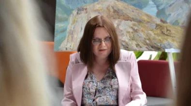 Artystka siedząca na wózku inwalidzkim przemawia podczas wernisażu wystawy w Mediatece 800-lecia. Ubrana w wzorzystą sukienkę i jasnoróżową marynarkę, trzyma w dłoniach kartki i patrzy w stronę publiczności. W tle obraz przedstawiający majestatyczne górskie krajobrazy z rzeką i kozicami, ustawiony na sztaludze. W dolnym rogu widoczne logo Mediateki.