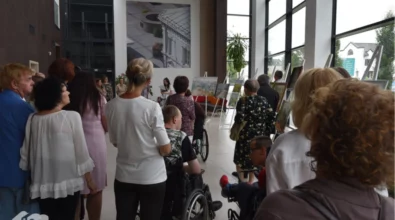 Wernisaż wystawy malarskiej w Mediatece 800-lecia. Liczna publiczność zgromadzona w jasnym, przeszklonym holu, gdzie na sztalugach prezentowane są obrazy. Wśród uczestników osoby starsze, kobiety i mężczyźni w eleganckich strojach, a także osoby poruszające się na wózkach inwalidzkich. W tle prowadząca wydarzenie kobieta odczytuje tekst, a za nią widać wielkoformatową fotografię architektoniczną na ścianie. W lewym dolnym rogu widoczne logo Mediateki.