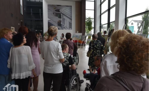 Wernisaż wystawy malarskiej w Mediatece 800-lecia. Liczna publiczność zgromadzona w jasnym, przeszklonym holu, gdzie na sztalugach prezentowane są obrazy. Wśród uczestników osoby starsze, kobiety i mężczyźni w eleganckich strojach, a także osoby poruszające się na wózkach inwalidzkich. W tle prowadząca wydarzenie kobieta odczytuje tekst, a za nią widać wielkoformatową fotografię architektoniczną na ścianie. W lewym dolnym rogu widoczne logo Mediateki.