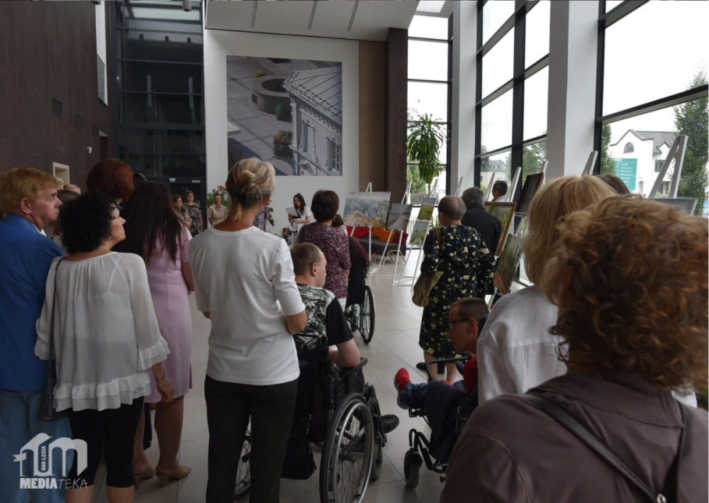 Wernisaż wystawy malarskiej w Mediatece 800-lecia. Liczna publiczność zgromadzona w jasnym, przeszklonym holu, gdzie na sztalugach prezentowane są obrazy. Wśród uczestników osoby starsze, kobiety i mężczyźni w eleganckich strojach, a także osoby poruszające się na wózkach inwalidzkich. W tle prowadząca wydarzenie kobieta odczytuje tekst, a za nią widać wielkoformatową fotografię architektoniczną na ścianie. W lewym dolnym rogu widoczne logo Mediateki.