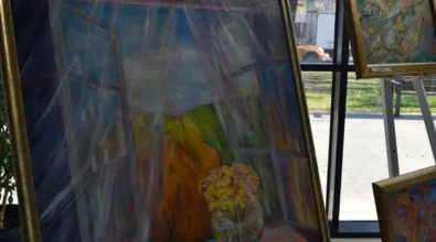 Oprawiony obraz malarski przedstawiający martwą naturę z bukietem kwiatów ustawionym przy oknie, utrzymany w stonowanej, wielobarwnej tonacji. Zdjęcie ukazuje fragment wystawy malarstwa Joanny Prus prezentowanej w przestrzeni Mediateki 800-lecia.