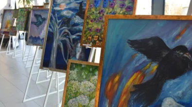 Na sztalugach ustawione są obrazy malarskie o żywej kolorystyce, przedstawiające motywy roślinne, krajobrazowe oraz symboliczne formy zwierzęce. Zdjęcie ukazuje fragment wystawy malarstwa Joanny Prus prezentowanej w przestrzeni Mediateki 800-lecia.