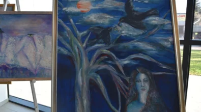 Oprawiony obraz malarski przedstawiający symboliczne połączenie pejzażu z kobiecą postacią i motywem ptaka na tle dynamicznego nieba. Zdjęcie ukazuje fragment wystawy malarstwa Joanny Prus prezentowanej w przestrzeni Mediateki 800-lecia.