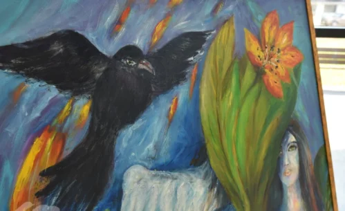 Fragment obrazu malarskiego przedstawiający czarnego ptaka z rozpostartymi skrzydłami na tle intensywnie kolorowanej, symbolicznej kompozycji z motywami roślinnymi i kobiecą twarzą. Zdjęcie ukazuje detal jednego z obrazów prezentowanych na wystawie malarstwa Joanny Prus w Mediatece 800-lecia.