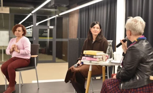 Joanna Kuciel-Frydryszak siedzi przy stoliku na scenie i rozmawia z prowadzącą spotkanie, która trzyma mikrofon. Na stoliku znajdują się książki autorki oraz karafka z wodą. Obok siedzi druga uczestniczka wydarzenia, która przysłuchuje się rozmowie.