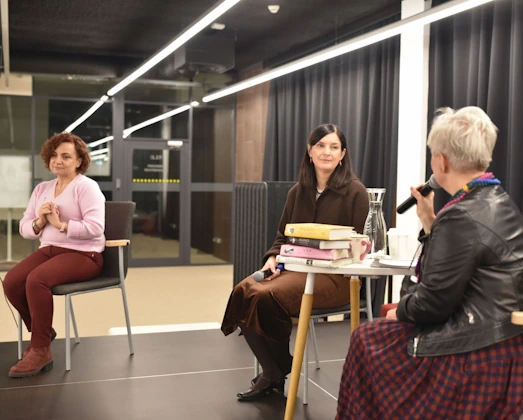 Joanna Kuciel-Frydryszak siedzi przy stoliku na scenie i rozmawia z prowadzącą spotkanie, która trzyma mikrofon. Na stoliku znajdują się książki autorki oraz karafka z wodą. Obok siedzi druga uczestniczka wydarzenia, która przysłuchuje się rozmowie.