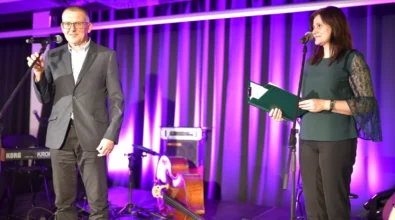 Mężczyzna stoi przy mikrofonie i przemawia do publiczności podczas wydarzenia na scenie. Obok niego stoi kobieta trzymająca zieloną teczkę z dokumentami. Tło sceny oświetlone jest fioletowym światłem, a za występującymi widoczne są instrumenty muzyczne oraz sprzęt nagłośnieniowy przygotowany do części artystycznej wydarzenia.