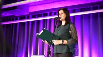 Kobieta stojąca przy mikrofonie przemawia do zgromadzonej publiczności, trzymając w rękach zieloną teczkę z przygotowanym tekstem wystąpienia. Scena oświetlona jest fioletowym światłem, które podkreśla uroczysty charakter wydarzenia. W tle widoczne są elementy nagłośnienia przygotowane do oprawy artystycznej spotkania.