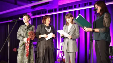 Cztery kobiety stoją na scenie podczas uroczystości odbywającej się w Mediatece. Jedna z nich czyta przygotowany tekst z zielonej teczki przy mikrofonie, a pozostałe trzymają dokumenty oraz bukiet kwiatów. Scena oświetlona jest fioletowym światłem, które podkreśla oficjalny charakter wydarzenia. W tle widoczne są instrumenty muzyczne przygotowane do oprawy artystycznej spotkania.