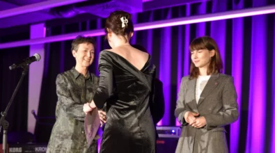 Trzy kobiety stoją na scenie. Jedna z nich wręcza dokument kobiecie ubranej w elegancką czarną suknię, a obok stoi kolejna uczestniczka wydarzenia. Scena oświetlona jest fioletowym światłem, a w tle widoczne są elementy nagłośnienia i instrumenty przygotowane do oprawy artystycznej wydarzenia.