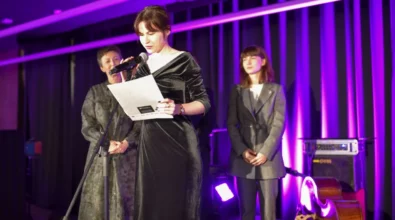 Kobieta stojąca przy mikrofonie odczytuje przygotowany tekst podczas oficjalnej części wydarzenia odbywającego się na scenie. Obok niej stoją dwie inne kobiety uczestniczące w uroczystości. Scena oświetlona jest dekoracyjnym fioletowym światłem, a w tle widoczne są elementy nagłośnienia oraz instrument muzyczny przygotowany do oprawy artystycznej wydarzenia.