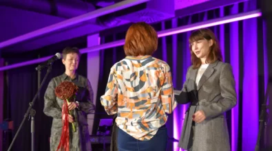 Na scenie trzy kobiety uczestniczą w uroczystym wręczeniu dokumentu. Jedna z kobiet w szarym garniturze przekazuje dokument stojącej przed nią osobie, widocznej tyłem, natomiast druga kobieta stojąca obok trzyma w rękach bukiet kwiatów przewiązany czerwoną wstążką. W tle widoczny jest mikrofon oraz scenografia oświetlona fioletowym światłem.