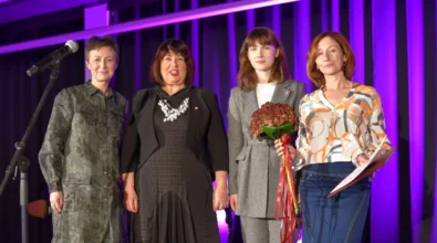 Cztery kobiety stoją na scenie i pozują do wspólnego zdjęcia po zakończeniu uroczystości. Jedna z nich trzyma bukiet kwiatów przewiązany czerwoną wstążką, a druga w dłoni trzyma dokument. Obok widoczny jest mikrofon, a tło sceny oświetlone jest intensywnym fioletowym światłem.