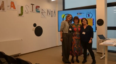 Zdjęcie prezentuje trzy kobiety pozujące do zdjęcia. Kobieta od lewej ma na sobie szare spodnie i bluzę, kobieta stojąca w środku kolorową sukienkę, a kobieta od prawej czarne spodnie i bluzkę. Z tyłu ekran monitora wyświetlający jakąś prezentację na niebieskim tle. Po lewej stronie widoczna biała ściana z napisem Majsternia. W lewym dolnym rogu zdjęcia widoczne czarne krzesła. Zdjęcie własnością MBP.