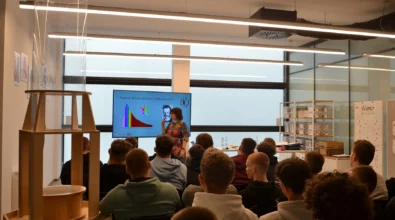 Zdjęcie prezentuje prawdopodobnie wykład. Na pierwszym planie grupa siedzących młodych osób widocznych od tyłu. Przed grupą stoi prezentująca coś kobieta. Ubrana w kolorową sukienkę. Za nią monitor z grafiką na niebieskim tle. Wydarzenie ma miejsce w niewielkim pomieszczeniu. Po lewej stronie widoczne wysokie drewniane urządzenie z przezroczystą tubą skierowaną ku górze. Zdjęcie własnością MBP.