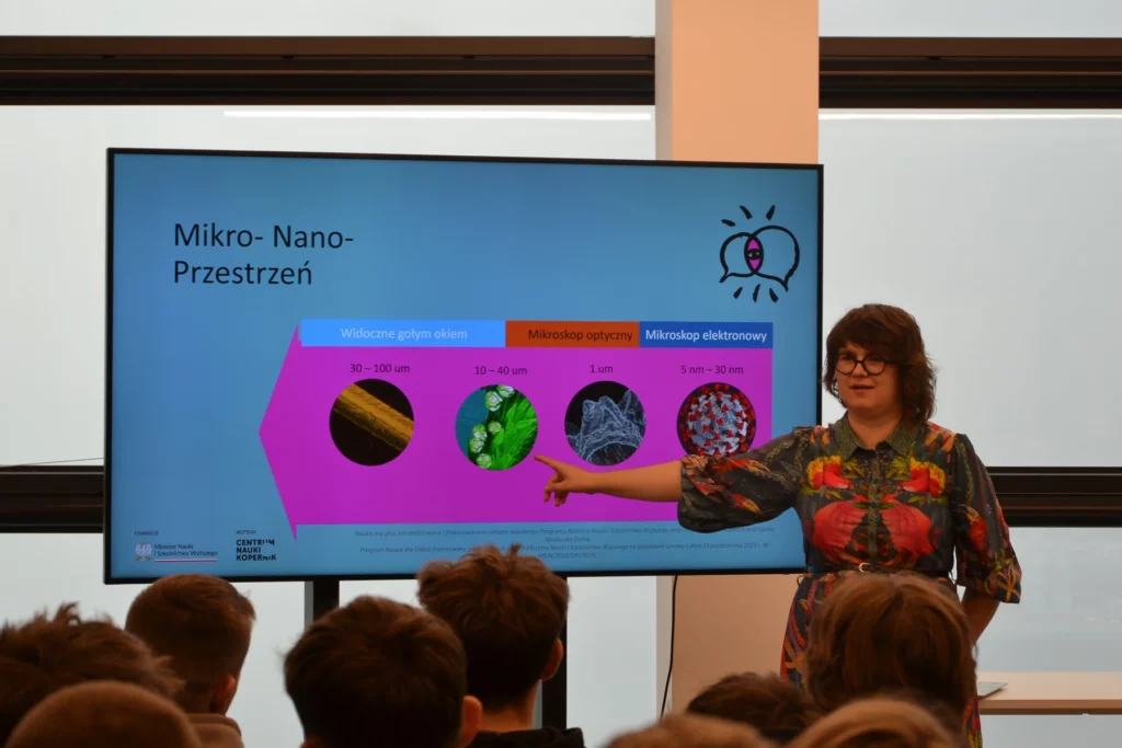 Zdjęcie prezentuje prawdopodobnie wykład. W centrum zdjęcia znajduje się stojąca osoba w kolorowej sukience. Kobieta ma krótkie włosy i okulary. Wyciągniętą ręką pokazuje na grafikę znajdującą się na monitorze. Obok niej widoczne są jeszcze trzy grafiki i opis. Prezentacja jest w kolorach niebieskim i różowym. Widoczny jest napis Nauka ma głos. Wydarzenie ma miejsce w niewielkim pomieszczeniu. Widać od tyłu siedzące młode osoby. Zdjęcie własnością MBP.