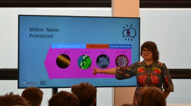 Zdjęcie prezentuje prawdopodobnie wykład. W centrum zdjęcia znajduje się stojąca osoba w kolorowej sukience. Kobieta ma krótkie włosy i okulary. Wyciągniętą ręką pokazuje na grafikę znajdującą się na monitorze. Obok niej widoczne są jeszcze trzy grafiki i opis. Prezentacja jest w kolorach niebieskim i różowym. Widoczny jest napis Nauka ma głos. Wydarzenie ma miejsce w niewielkim pomieszczeniu. Widać od tyłu siedzące młode osoby. Zdjęcie własnością MBP.