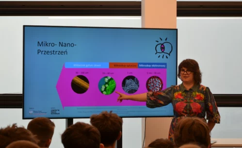 Zdjęcie prezentuje prawdopodobnie wykład. W centrum zdjęcia znajduje się stojąca osoba w kolorowej sukience. Kobieta ma krótkie włosy i okulary. Wyciągniętą ręką pokazuje na grafikę znajdującą się na monitorze. Obok niej widoczne są jeszcze trzy grafiki i opis. Prezentacja jest w kolorach niebieskim i różowym. Widoczny jest napis Nauka ma głos. Wydarzenie ma miejsce w niewielkim pomieszczeniu. Widać od tyłu siedzące młode osoby. Zdjęcie własnością MBP.