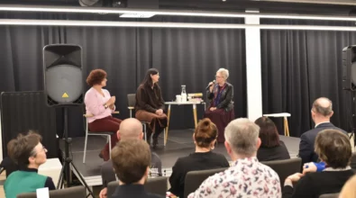 Na scenie Mediateki 800-lecia odbywa się spotkanie autorskie z Joanną Kuciel-Frydryszak. Autorka siedzi przy stoliku z książkami i rozmawia z prowadzącą wydarzenie, która trzyma mikrofon. Po lewej stronie obecna jest tłumaczka języka migowego, przekładająca przebieg rozmowy dla uczestników. W pierwszych rzędach siedzi publiczność uważnie słuchająca dyskusji literackiej.