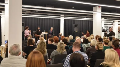 Publiczność siedzi w rzędach krzeseł i słucha spotkania autorskiego odbywającego się w Mediatece 800-lecia. Na scenie Joanna Kuciel-Frydryszak prowadzi rozmowę z zaproszonymi gościniami. Jedna z uczestniczek trzyma mikrofon i odpowiada na pytania podczas dyskusji. Z boku sali widoczna jest osoba tłumacząca wypowiedzi na język migowy oraz operator kamery rejestrujący przebieg wydarzenia.