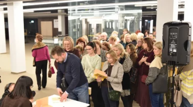 Uczestnicy spotkania ustawili się w kolejce, aby zdobyć autograf Joanny Kuciel-Frydryszak. Autorka siedzi przy stoliku i podpisuje książki przekazywane przez czytelników. W kolejce stoją osoby trzymające egzemplarze publikacji, oczekujące na możliwość krótkiej rozmowy i dedykacji.