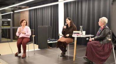 Joanna Kuciel-Frydryszak siedzi przy stoliku na scenie i rozmawia z prowadzącą spotkanie, która trzyma mikrofon. Na stoliku znajdują się książki autorki oraz karafka z wodą. Obok siedzi druga uczestniczka wydarzenia, która przysłuchuje się rozmowie.