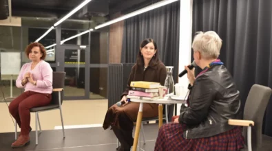 Joanna Kuciel-Frydryszak siedzi na scenie i rozmawia z prowadzącą spotkanie, trzymając mikrofon w dłoni. Na stoliku obok znajdują się książki autorki, karafka z wodą oraz kubek. Po lewej stronie siedzi tłumaczka języka migowego, która przekłada rozmowę dla uczestników wydarzenia.