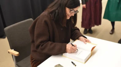 Joanna Kuciel-Frydryszak siedzi przy stoliku i podpisuje egzemplarz swojej książki „Chłopki”. Autorka pochyla się nad otwartą publikacją, wpisując dedykację dla czytelnika. Na stoliku leżą długopisy oraz osobisty stempel, którym oznaczane są podpisywane książki.