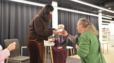 Joanna Kuciel-Frydryszak stoi na scenie i odbiera od uczestnika spotkania kartkę lub notatkę. Mężczyzna stojący przed sceną trzyma mikrofon i przekazuje autorce przygotowaną wiadomość. W tle siedzi prowadząca spotkanie, a na stoliku obok znajdują się książki autorki.