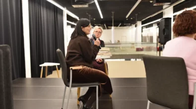 Joanna Kuciel-Frydryszak siedzi na scenie i trzyma mikrofon, zabierając głos podczas spotkania autorskiego. Obok niej siedzi uczestniczka rozmowy, która słucha wypowiedzi autorki. Na stoliku znajdują się książki oraz karafka z wodą. Wydarzenie odbywa się w przestrzeni Mediateki 800-lecia w Piotrkowie Trybunalskim.